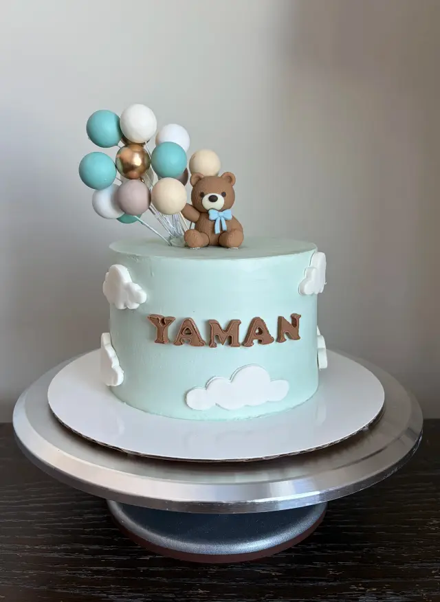 Pale blue cake with a teddy bear topper, a cluster of balloons and a nameplate reading Yaman.