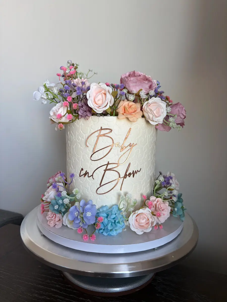 A white buttercream cake covered in pastel sugar flowers with gold 'Baby in Bloom' lettering, on a silver cake stand.