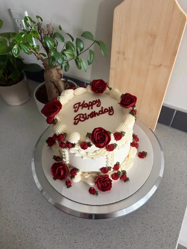 Ivory buttercream cake with piped shell borders, deep red buttercream roses and buds, and Happy Birthday in red script on top.