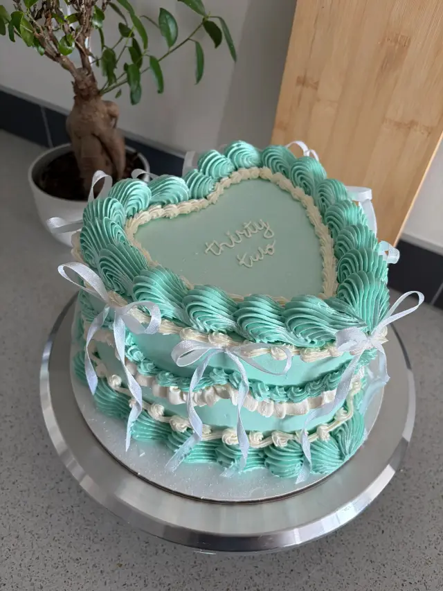 Mint green heart-shaped cake with white vintage-style piping around the edges and a white ribbon bow.