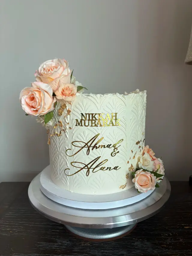 Tall ivory buttercream cake trimmed with peach and cream sugar roses, with Nikkah Mubarak in gold script.