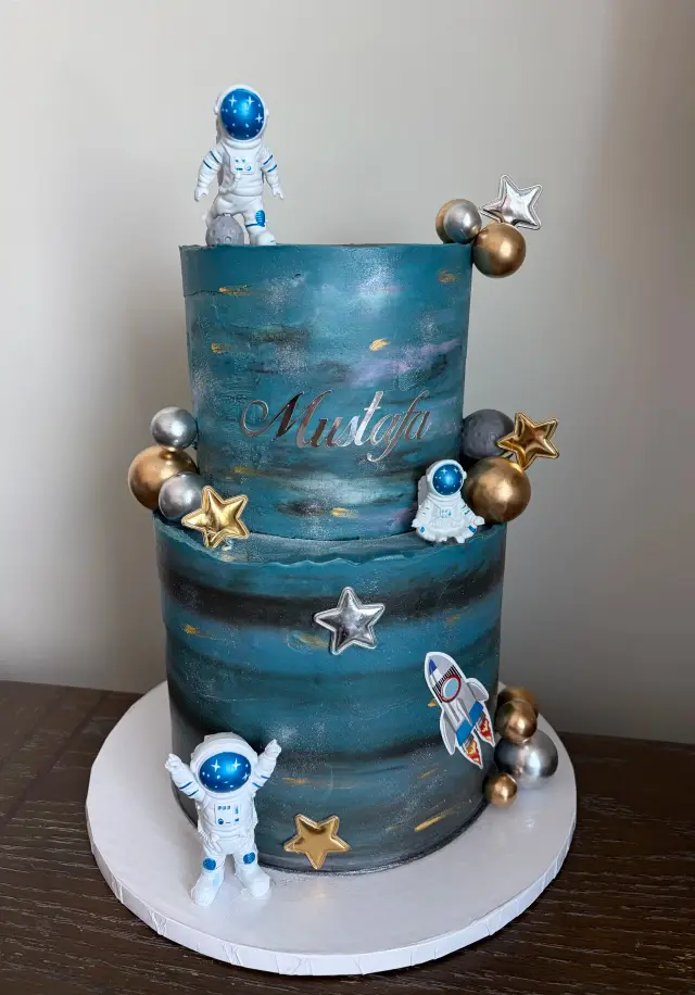 Tall navy cake painted like a night sky with gold stars and planets, and an astronaut figure on top.