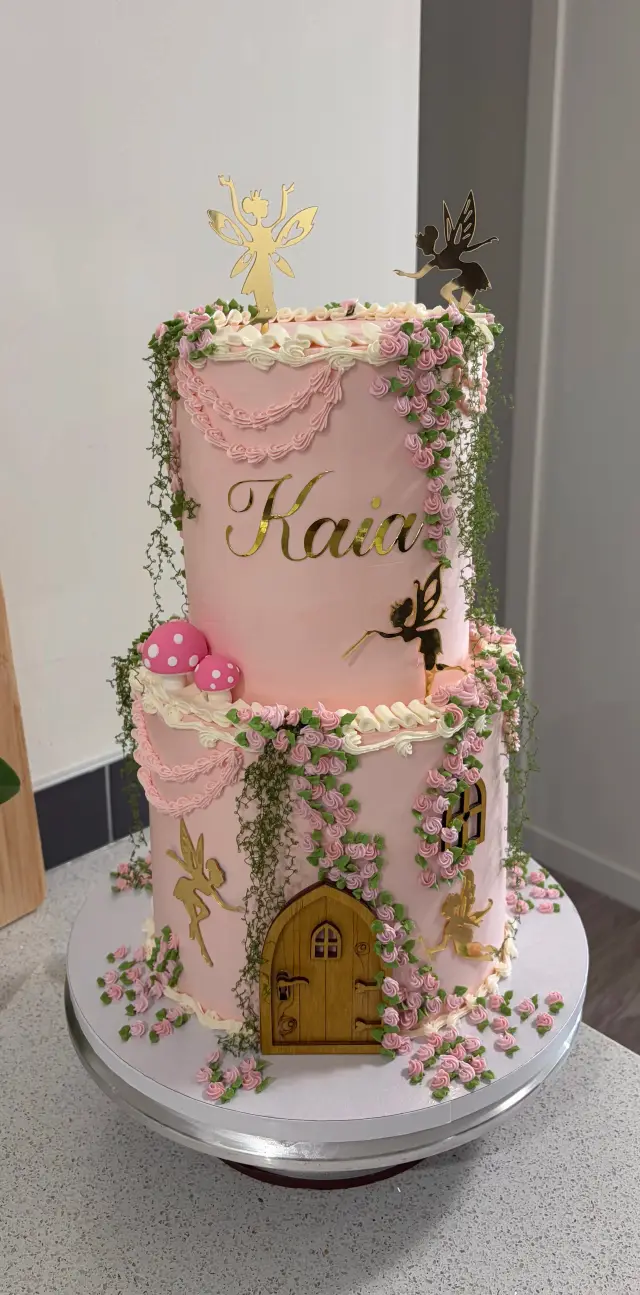 Two tier pink and cream cake decorated as a fairy garden, with a tiny door, climbing flowers and a gold topper.