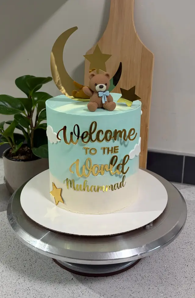Pale blue and cream ombre cake with white fondant clouds, gold stars and a moon, a teddy bear topper, and Welcome to the World Muhammad in gold lettering.