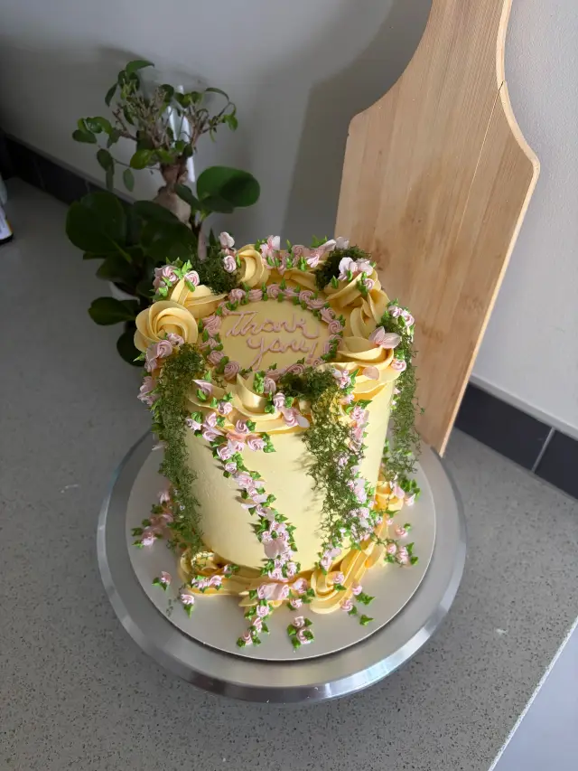 Tall butter yellow cake with piped swirls, trailing pink blossoms and green ivy down the sides, and Thank you in pink script on top.